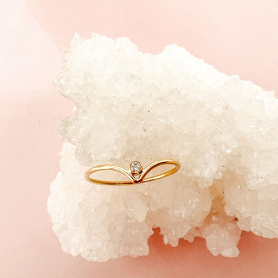 14K gold filled chevron shaped ring with white sparkly cubic Zirconia stone nestled into the centre point of the chevron peak. Ring is displayed onto of a white sparkly crystal geode with a pink background 
