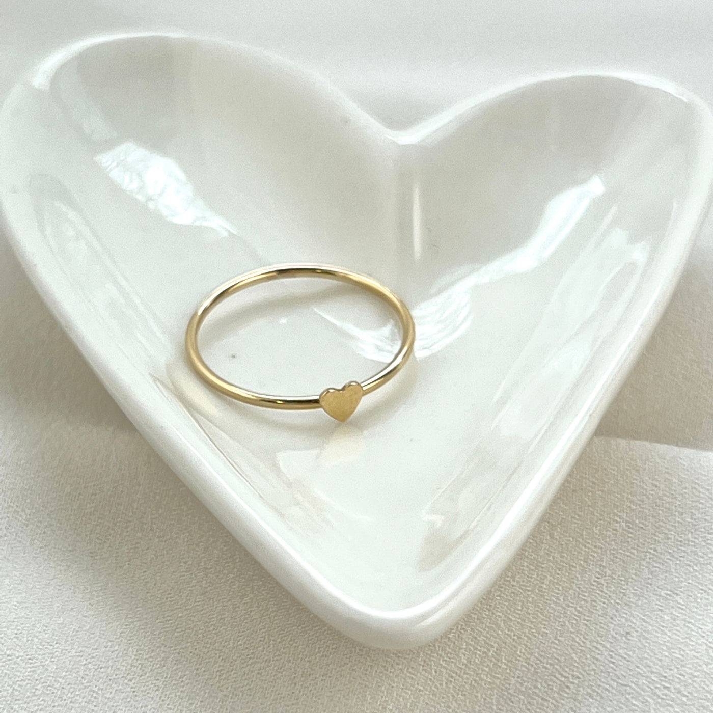 14K Gold filled simple band ring with tiny 2mm cut out heart shape on top. Gold heart ring is in a white shiny heart shaped jewelry dish