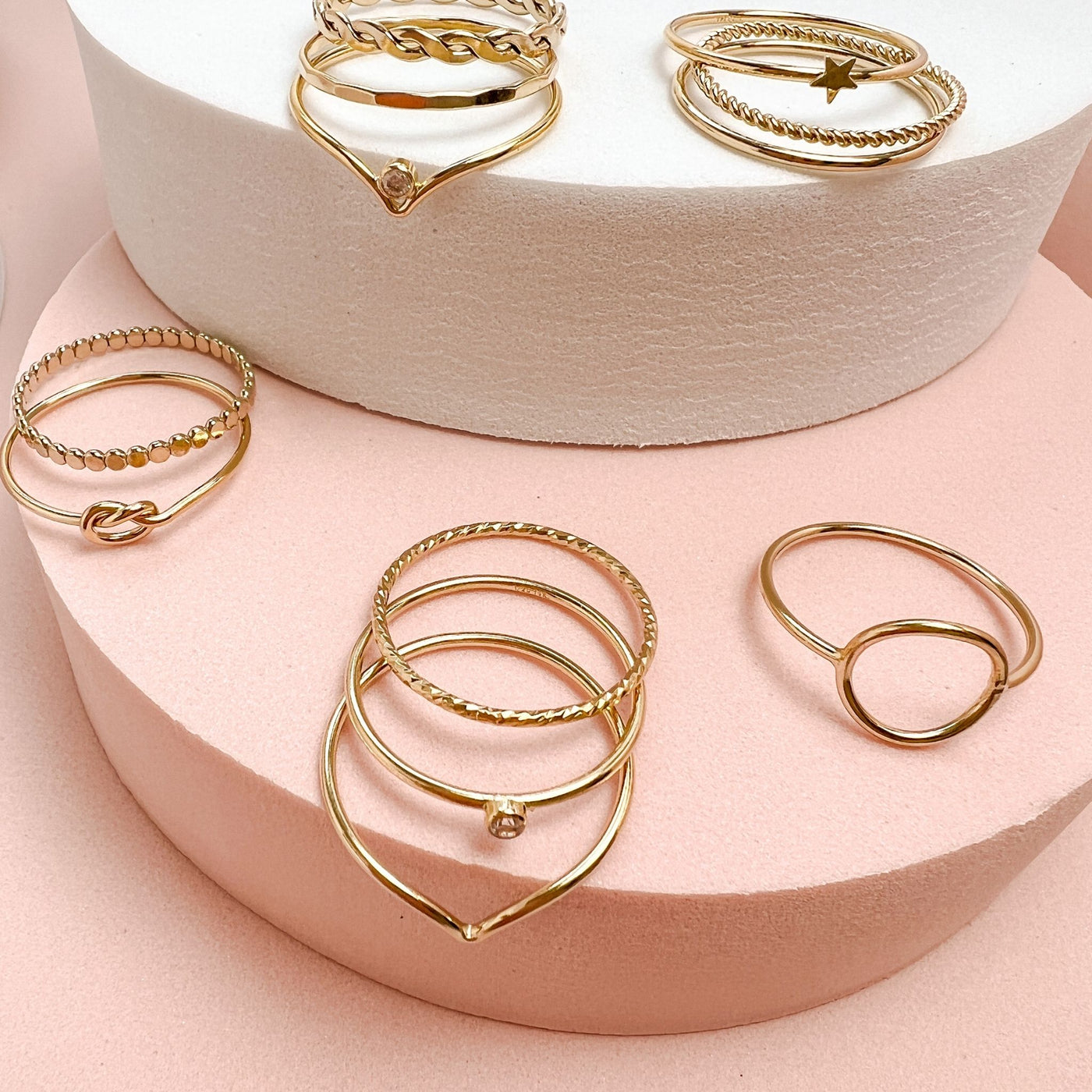 Assortment of differing styles of minimalist and dainty women’s stacking rings displayed on pink and white coloured round displays